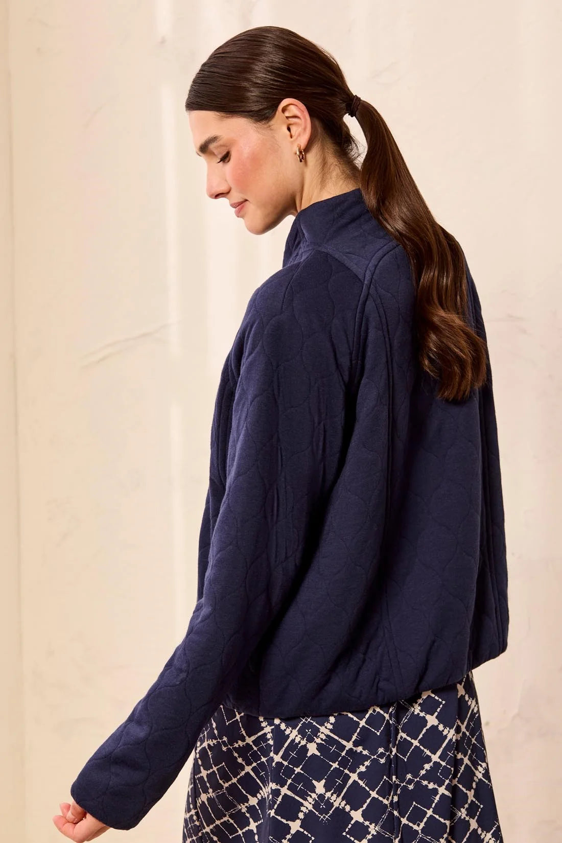 A woman wearing a dark navy blue textured long-sleeve jacket and a patterned skirt, shown from a side-back angle against a light beige wall.