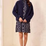 A woman modeling a navy blue zip-up jacket over a navy and white patterned outfit, standing in a minimalist cream-colored room with light wood floors.