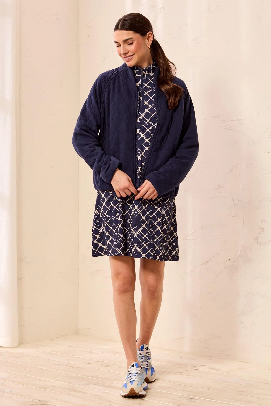 A woman modeling a navy blue zip-up jacket over a navy and white patterned outfit, standing in a minimalist cream-colored room with light wood floors.