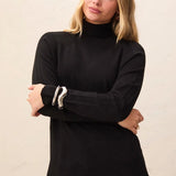 A woman wearing a black long-sleeve mock neck top with silver metallic cuff accents, paired with gray jeans, against a plain beige background.