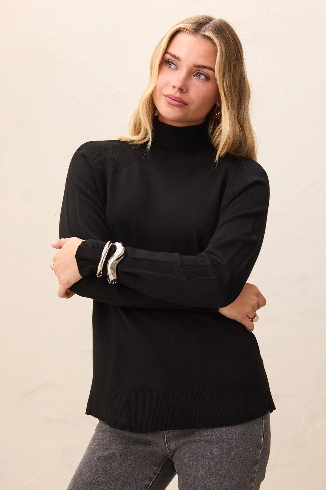 A woman wearing a black long-sleeve mock neck top with silver metallic cuff accents, paired with gray jeans, against a plain beige background.