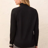 A woman shown from the back wearing a black long-sleeve mock neck top and gray pants, standing against a textured off-white wall.