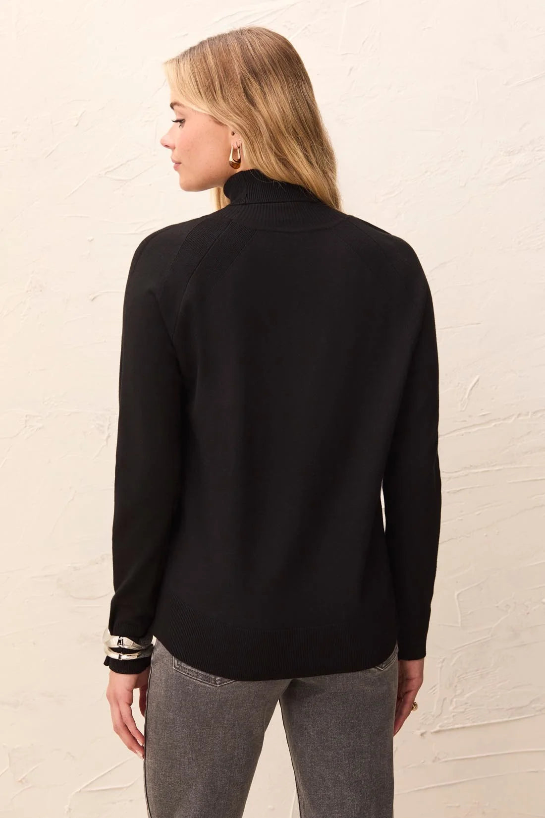 A woman shown from the back wearing a black long-sleeve mock neck top and gray pants, standing against a textured off-white wall.