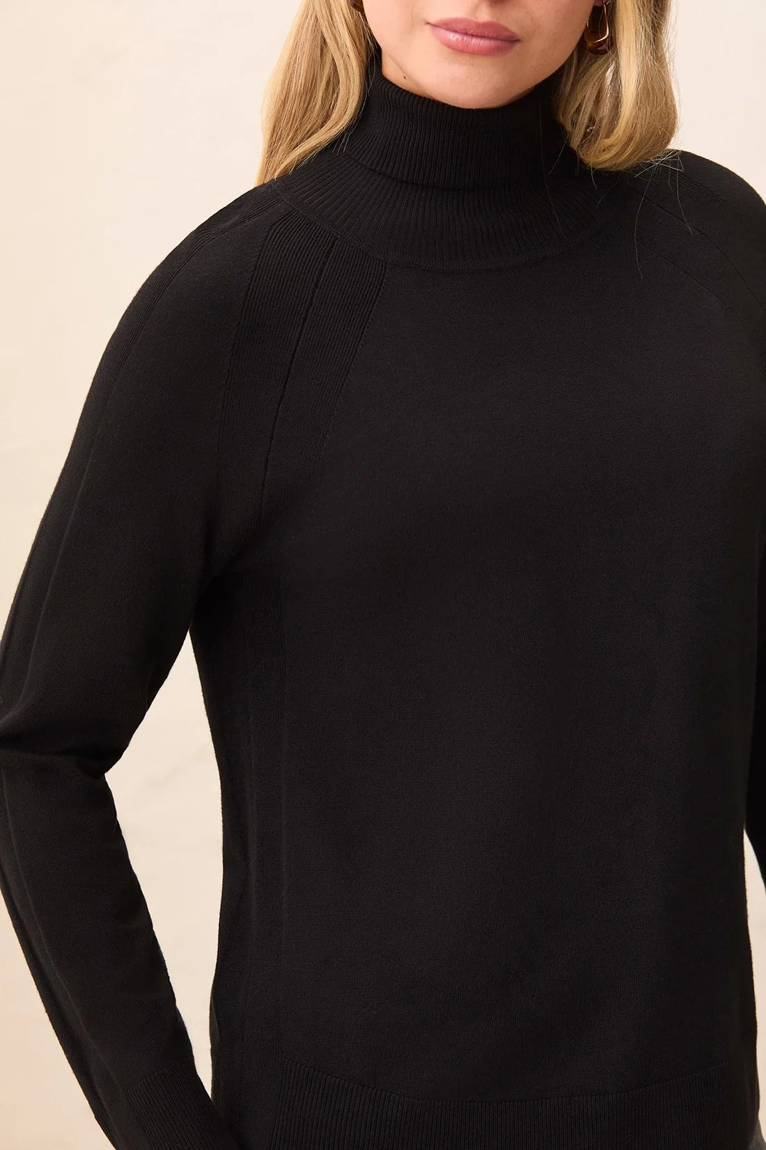 A close-up of a woman wearing a black ribbed knit turtleneck sweater against a light beige background.