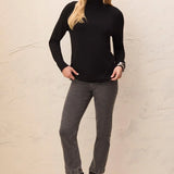 A woman modeling a black long-sleeve mock neck turtleneck sweater, paired with dark gray cropped jeans and black ankle boots, standing against a textured beige wall.