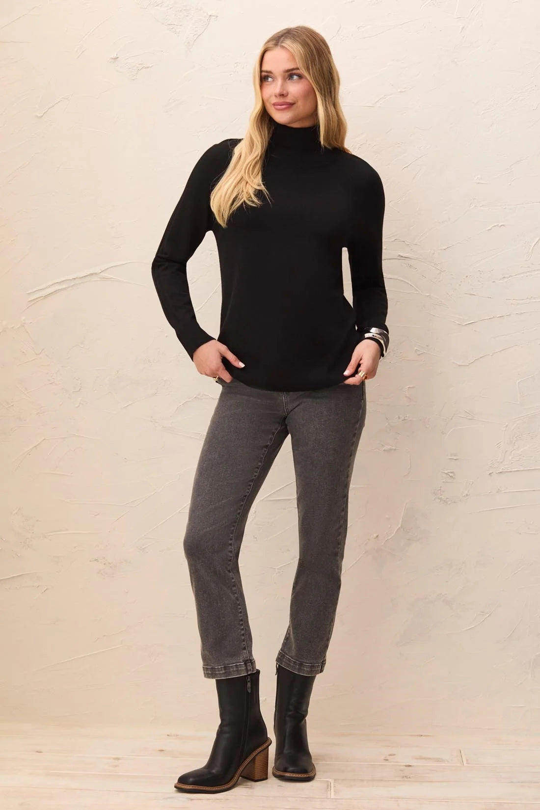 A woman modeling a black long-sleeve mock neck turtleneck sweater, paired with dark gray cropped jeans and black ankle boots, standing against a textured beige wall.