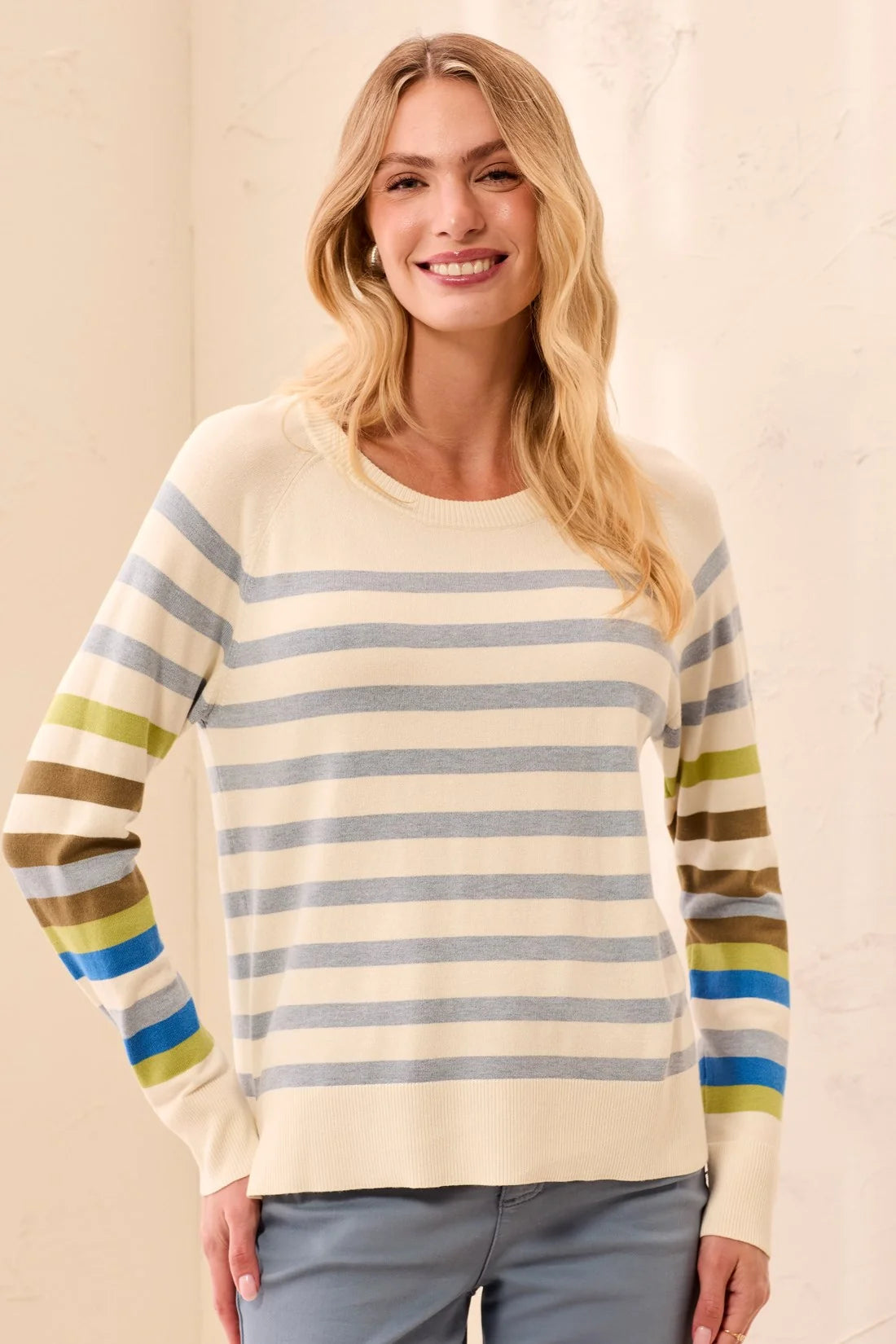 A woman wearing a cream-colored knit sweater with horizontal stripes in blue, olive green, and brown on the sleeves and body, paired with gray pants.