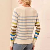 A woman shown from the back wearing a cream-colored knit sweater with horizontal stripes in blue, olive green, brown, and yellow on the sleeves, paired with light blue pants.
