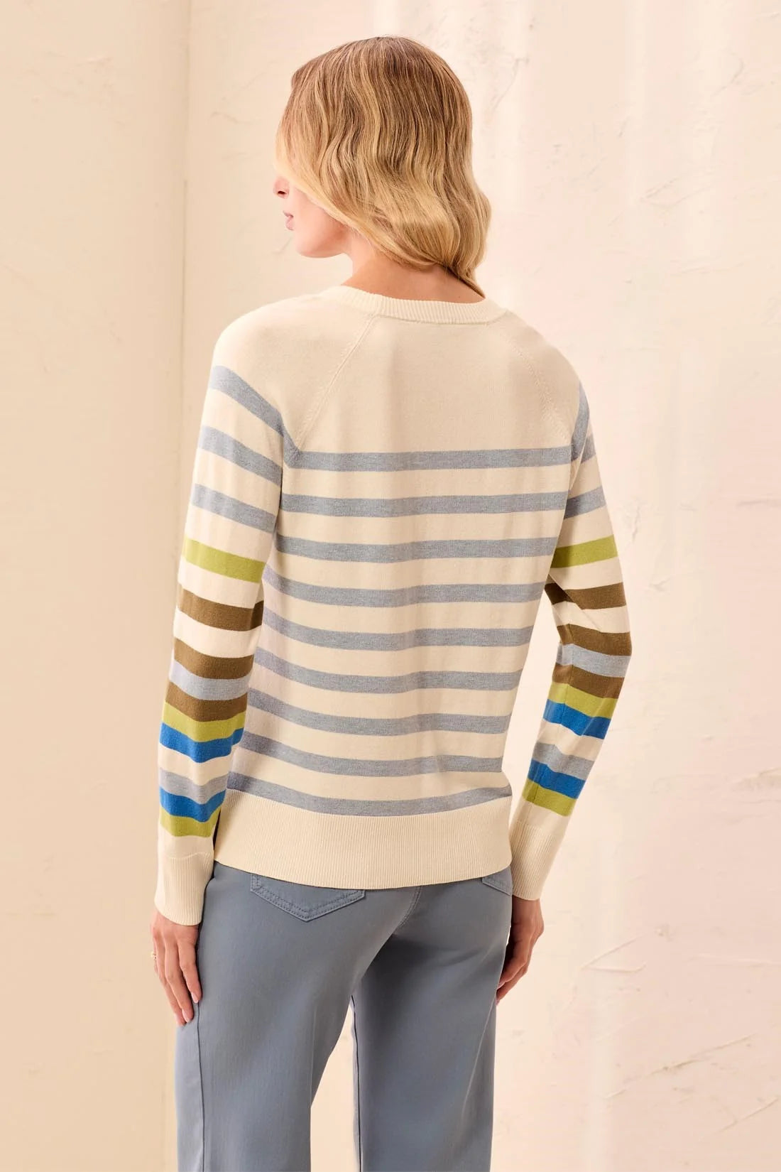 A woman shown from the back wearing a cream-colored knit sweater with horizontal stripes in blue, olive green, brown, and yellow on the sleeves, paired with light blue pants.