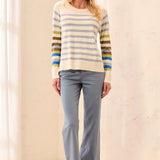 A woman modeling a cream knit sweater with horizontal blue, gray, olive green, and yellow stripes on the sleeves, paired with light blue pants and brown suede shoes in a bright indoor setting.