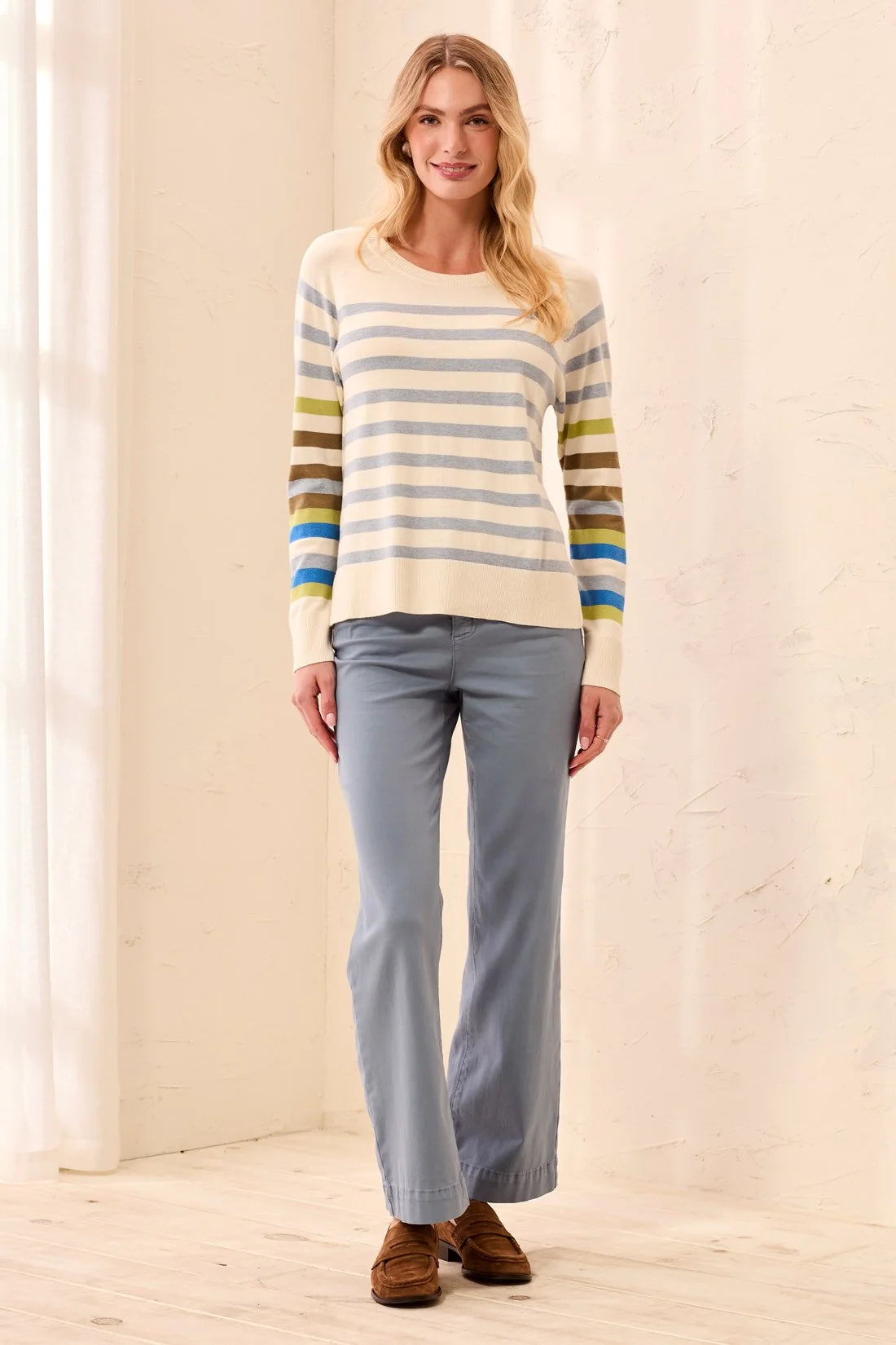 A woman modeling a cream knit sweater with horizontal blue, gray, olive green, and yellow stripes on the sleeves, paired with light blue pants and brown suede shoes in a bright indoor setting.