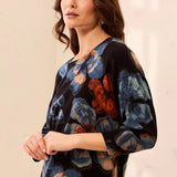 A woman wearing a black three-quarter sleeve blouse with a blue and rust-orange floral print, paired with gray jeans, standing against a light beige background.