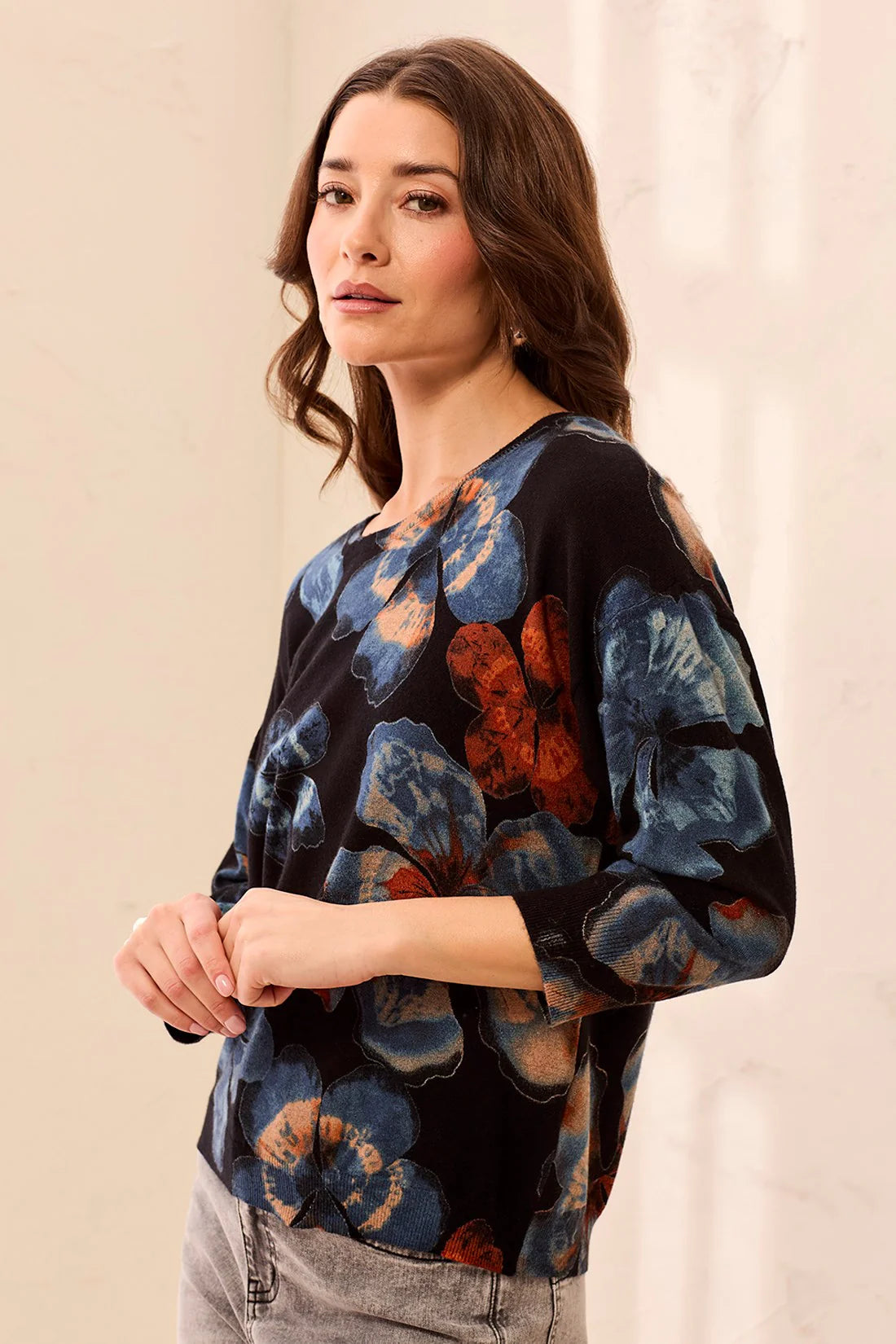 A woman wearing a black three-quarter sleeve blouse with a blue and rust-orange floral print, paired with gray jeans, standing against a light beige background.