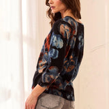 A woman wearing a black three-quarter sleeve blouse with a blue, orange, and brown floral pattern, paired with gray jeans, standing in a brightly lit room.