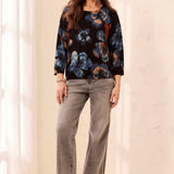 A woman modeling a black long-sleeve top with a blue and orange floral pattern, paired with gray cuffed jeans and black ankle boots, standing in a bright, neutral indoor setting.