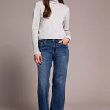 Woman wearing a light gray turtleneck sweater and blue jeans against a plain background