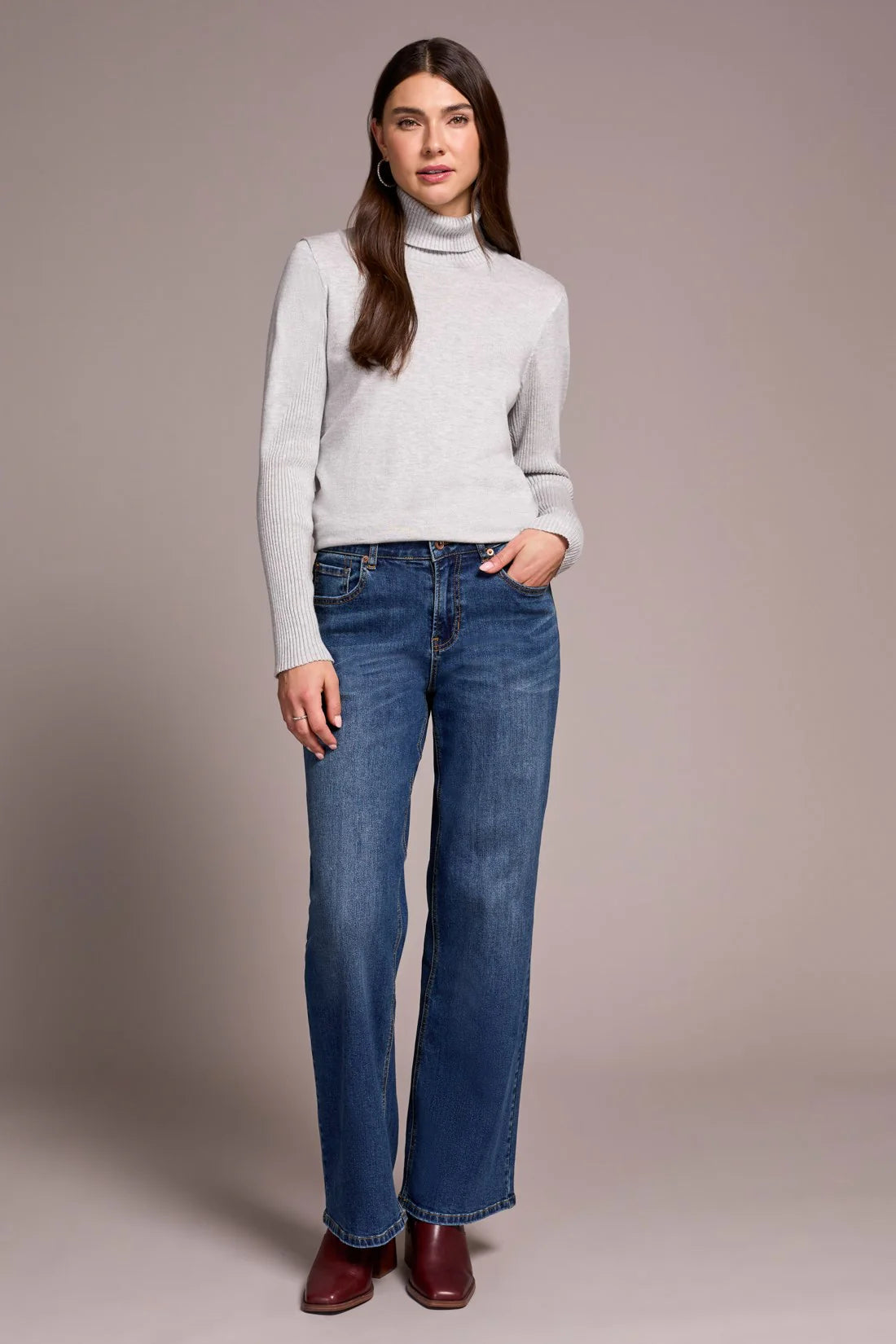 Woman wearing a light gray turtleneck sweater and blue jeans against a plain background