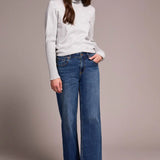 Woman wearing a white turtleneck sweater and blue jeans against a plain background