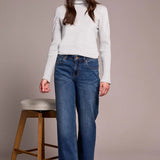 Woman wearing a white turtleneck sweater and blue jeans standing next to a wooden stool against a plain background.