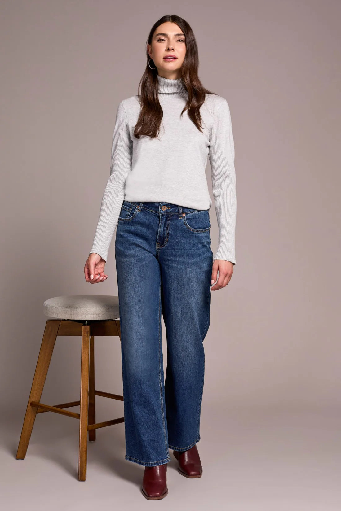 Woman wearing a white turtleneck sweater and blue jeans standing next to a wooden stool against a plain background.