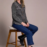 Woman sitting on a stool wearing a gray patterned shirt, blue jeans, and black boots against a plain background