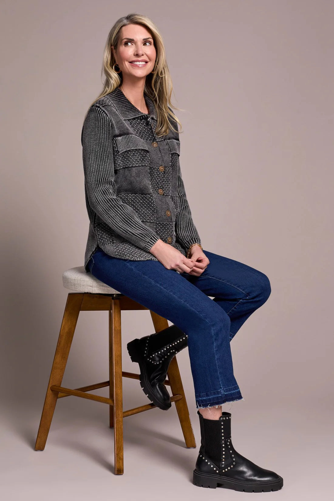 Woman sitting on a stool wearing a gray patterned shirt, blue jeans, and black boots against a plain background