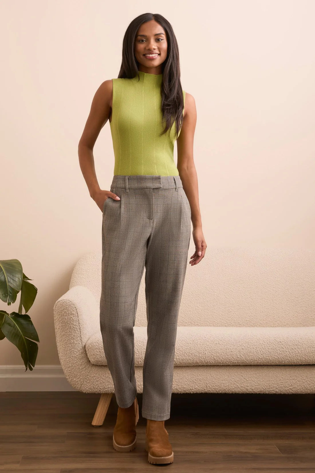 Woman wearing a green sleeveless top and gray checkered pants standing in a room with a beige sofa and plant.