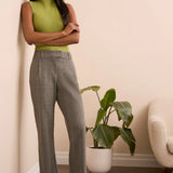 Woman in green top and gray checkered pants standing in a room with a plant and chair.