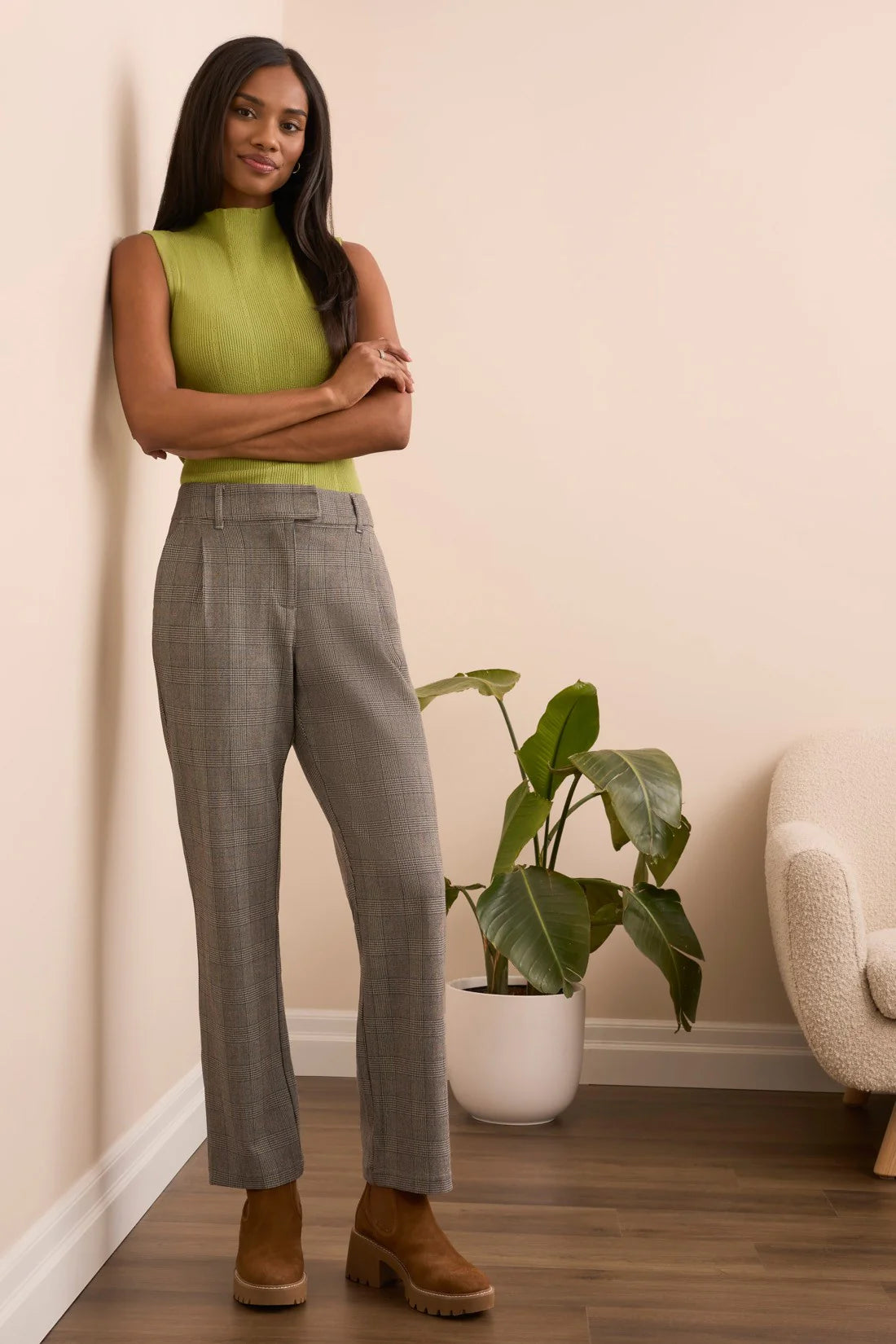 Woman in green top and gray checkered pants standing in a room with a plant and chair.