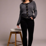 Woman wearing a gray sweater with black buttons and black pants standing next to a wooden stool on a plain background