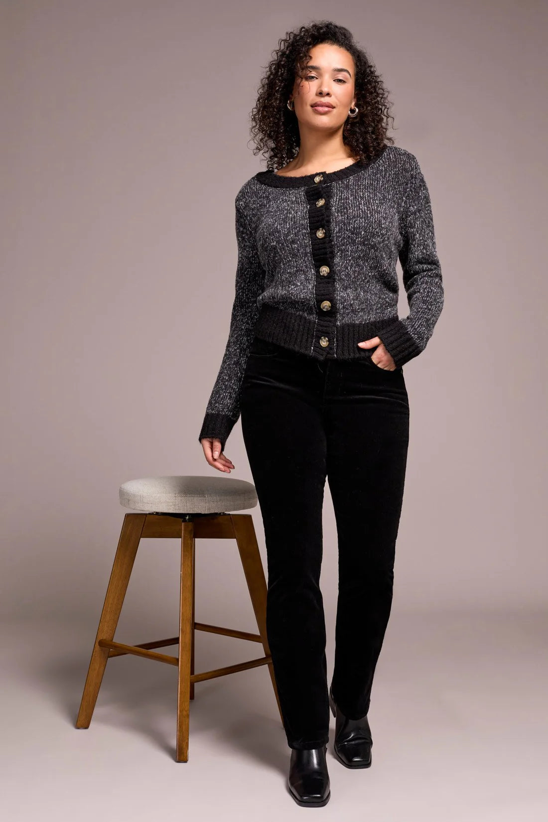 Woman wearing a gray sweater with black buttons and black pants standing next to a wooden stool on a plain background