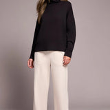 Woman wearing a black turtleneck and white pants against a gray background
