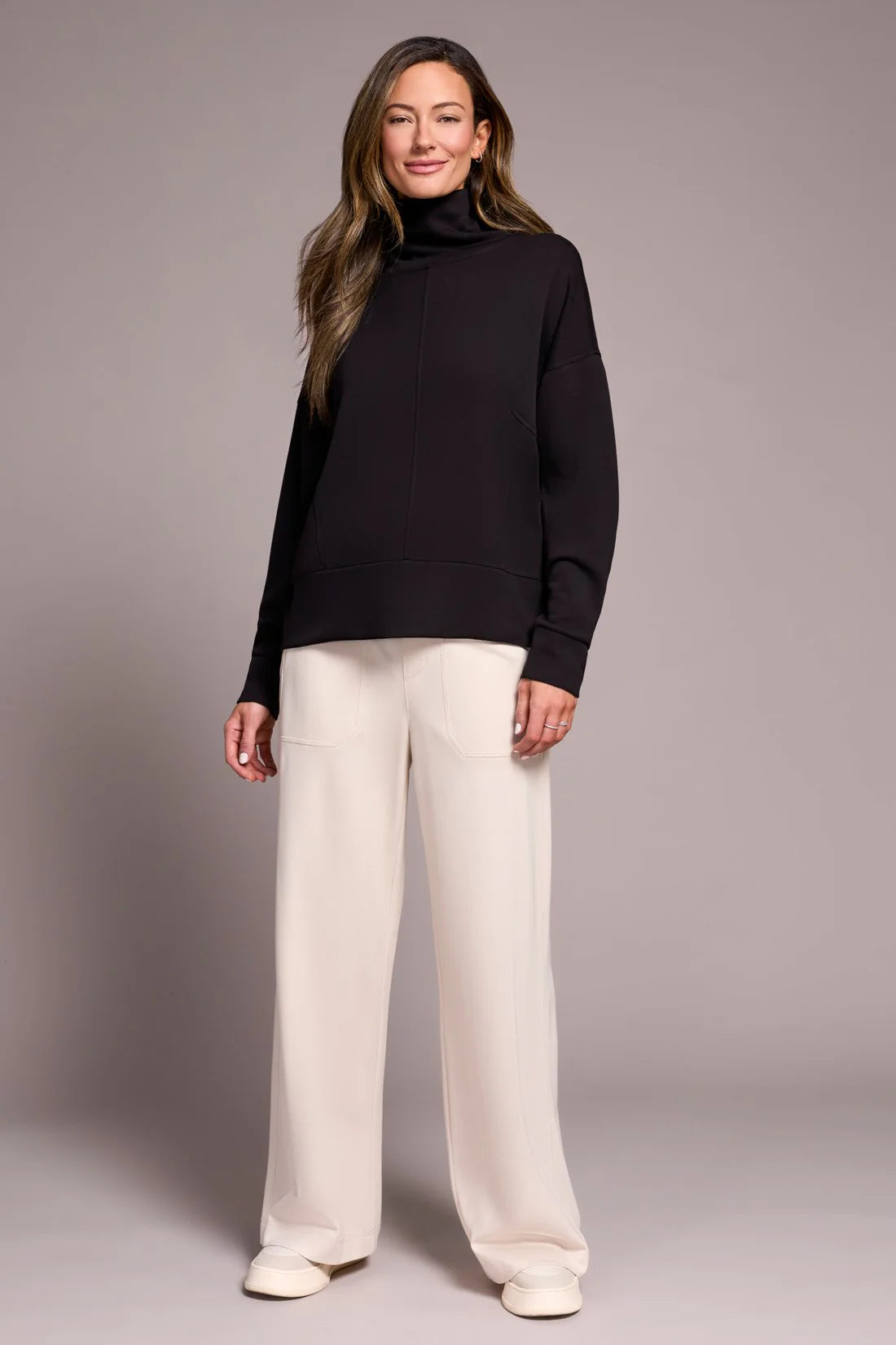 Woman wearing a black turtleneck and white pants against a gray background