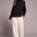 Woman wearing a black turtleneck and white pants against a gray background