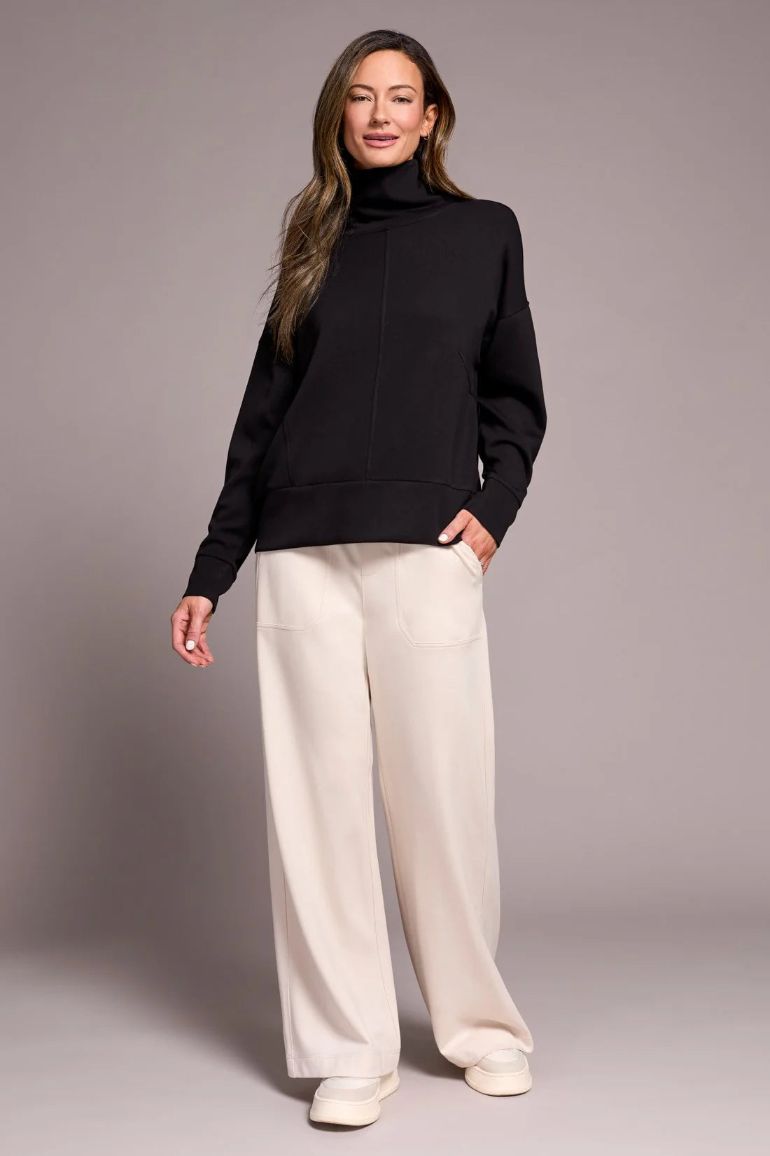 Woman wearing a black turtleneck and white pants against a gray background