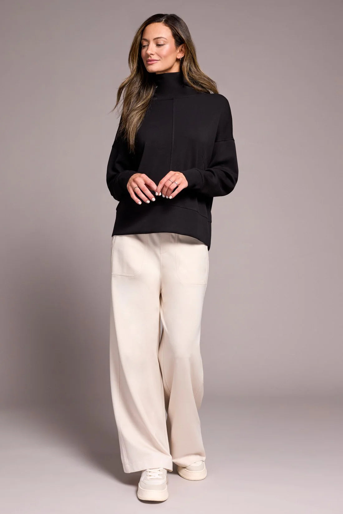Woman wearing a black turtleneck and white pants against a gray background
