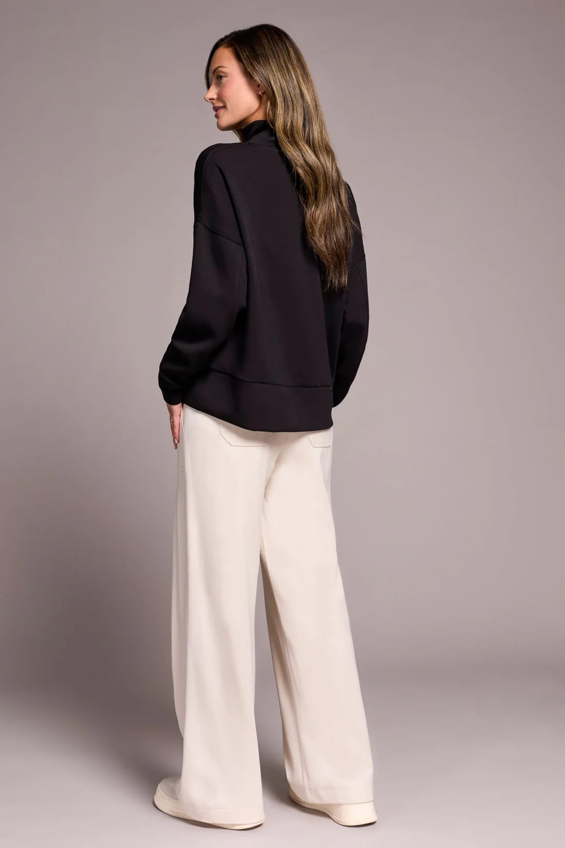 Woman wearing a black jacket and white pants against a plain background