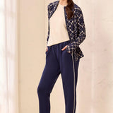 A woman models a navy blue two-piece lounge set consisting of a patterned zip-up jacket and solid navy pants with a white side stripe, styled with a cream top and beige sneakers.