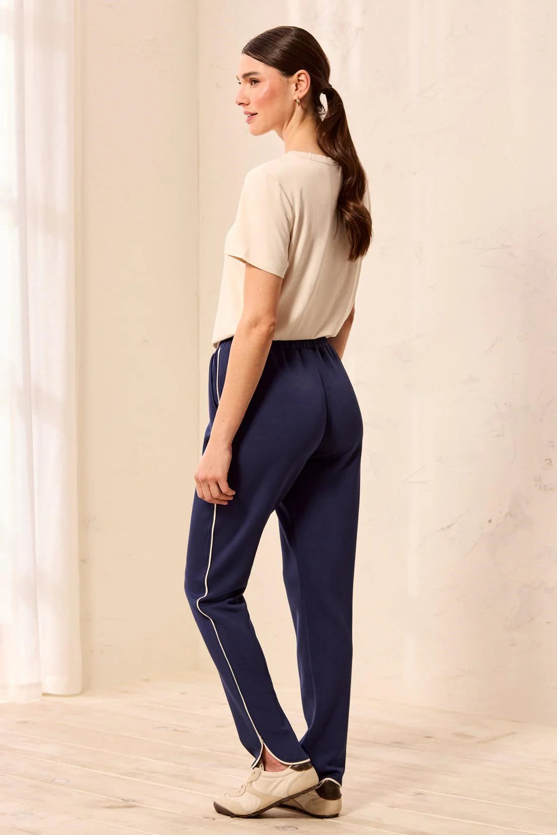 A woman wearing navy blue athletic pants with a thin white side stripe, paired with a beige t-shirt and beige sneakers, standing in a bright indoor setting.