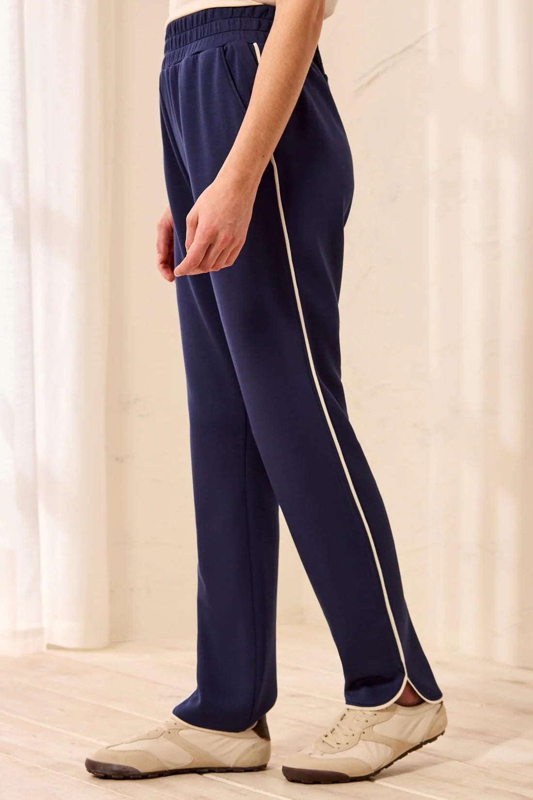 A person wearing navy blue athletic pants with a cream-colored side stripe, paired with beige sneakers, standing in a brightly lit indoor setting.