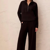 A woman modeling a dark brown matching loungewear set consisting of a half-zip long-sleeve top and wide-leg pants, paired with tan slip-on shoes, standing against a textured beige wall.
