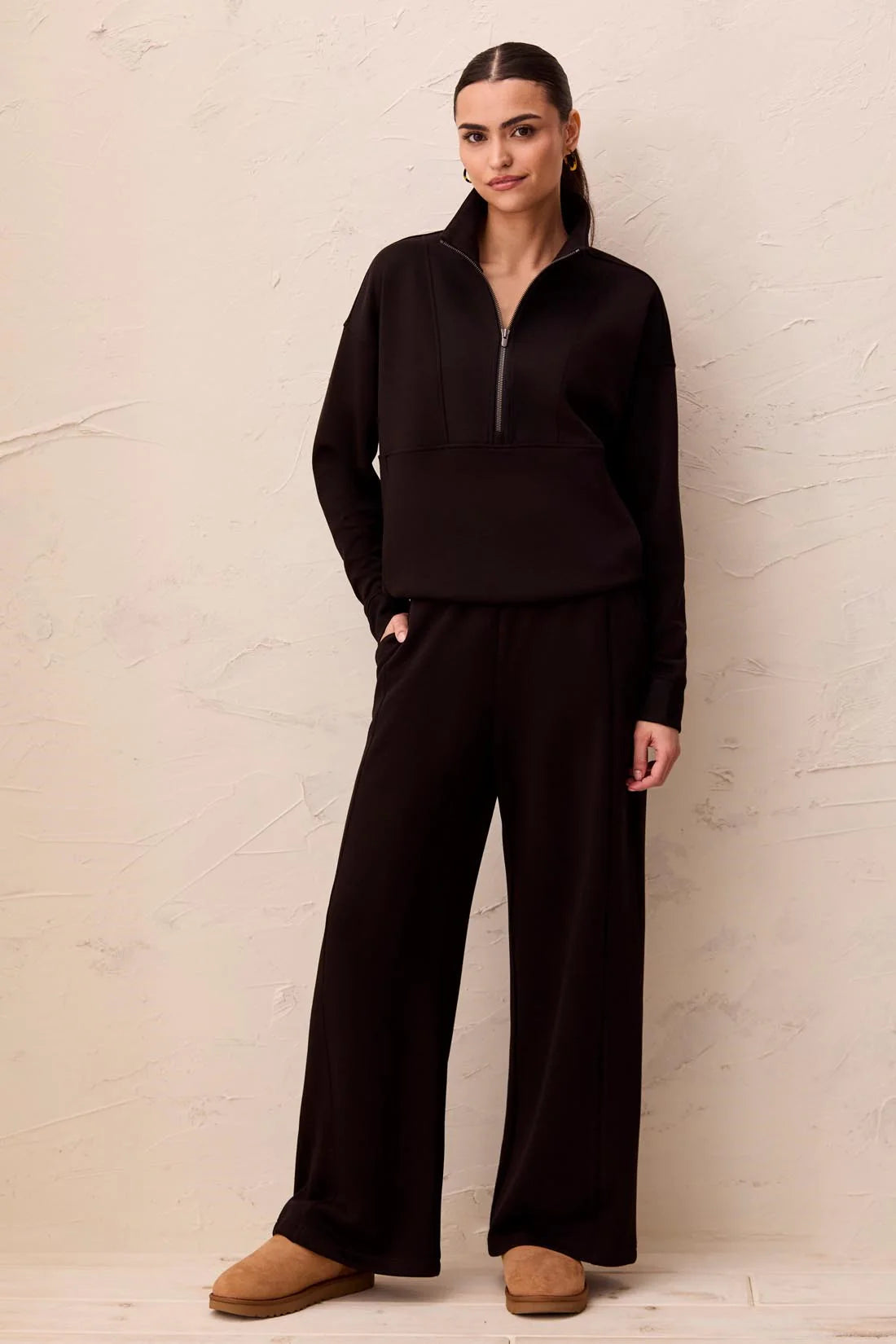 A woman modeling a dark brown matching loungewear set consisting of a half-zip long-sleeve top and wide-leg pants, paired with tan slip-on shoes, standing against a textured beige wall.