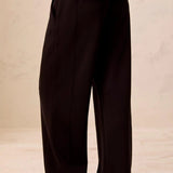 Black wide-leg trousers shown from the back on a model, paired with tan slip-on shoes against a neutral beige background.