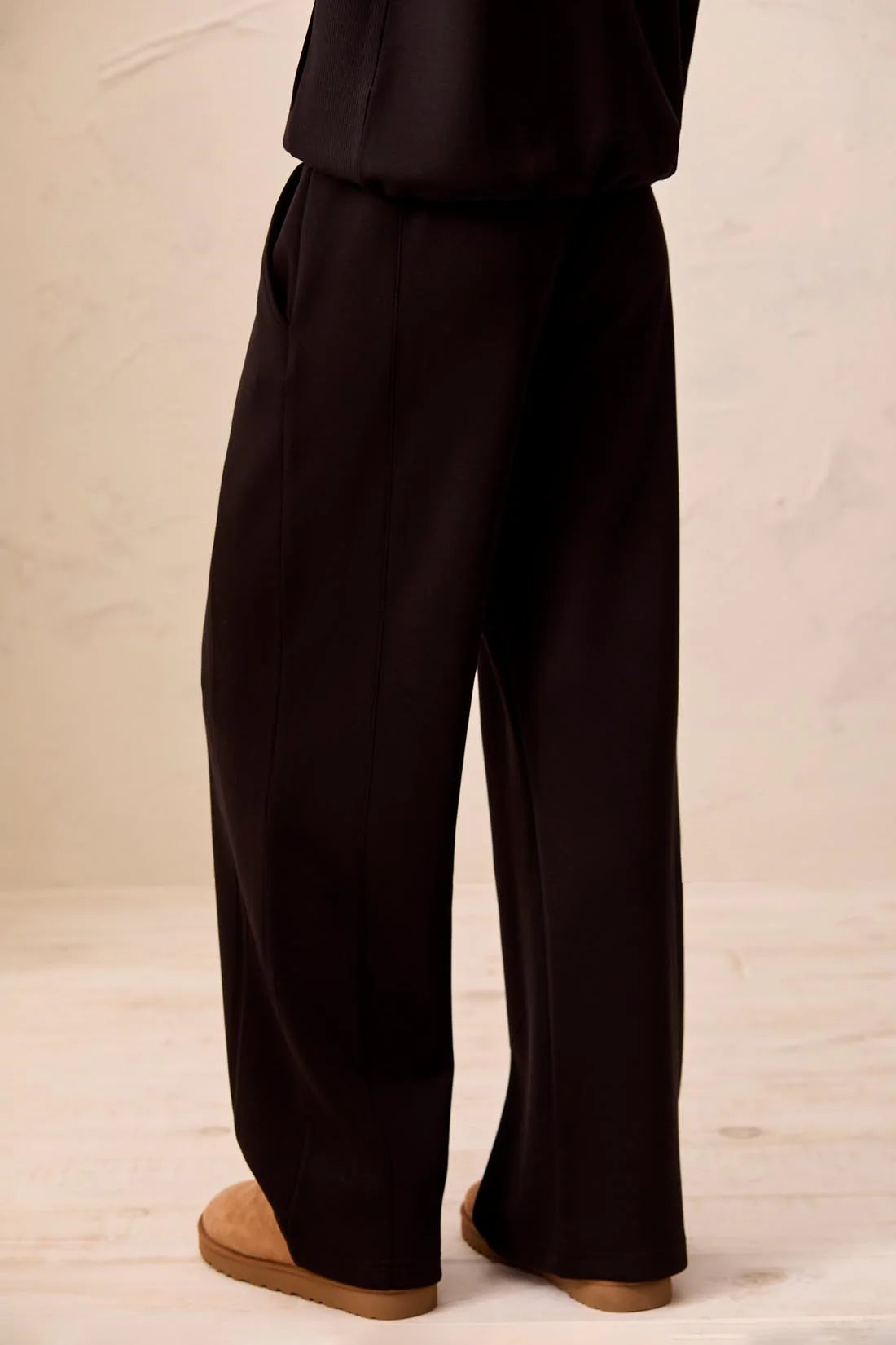 Black wide-leg trousers shown from the back on a model, paired with tan slip-on shoes against a neutral beige background.
