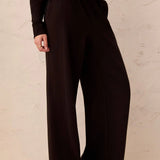 A person wearing dark brown wide-leg lounge pants with a matching dark top, standing against a textured beige wall.