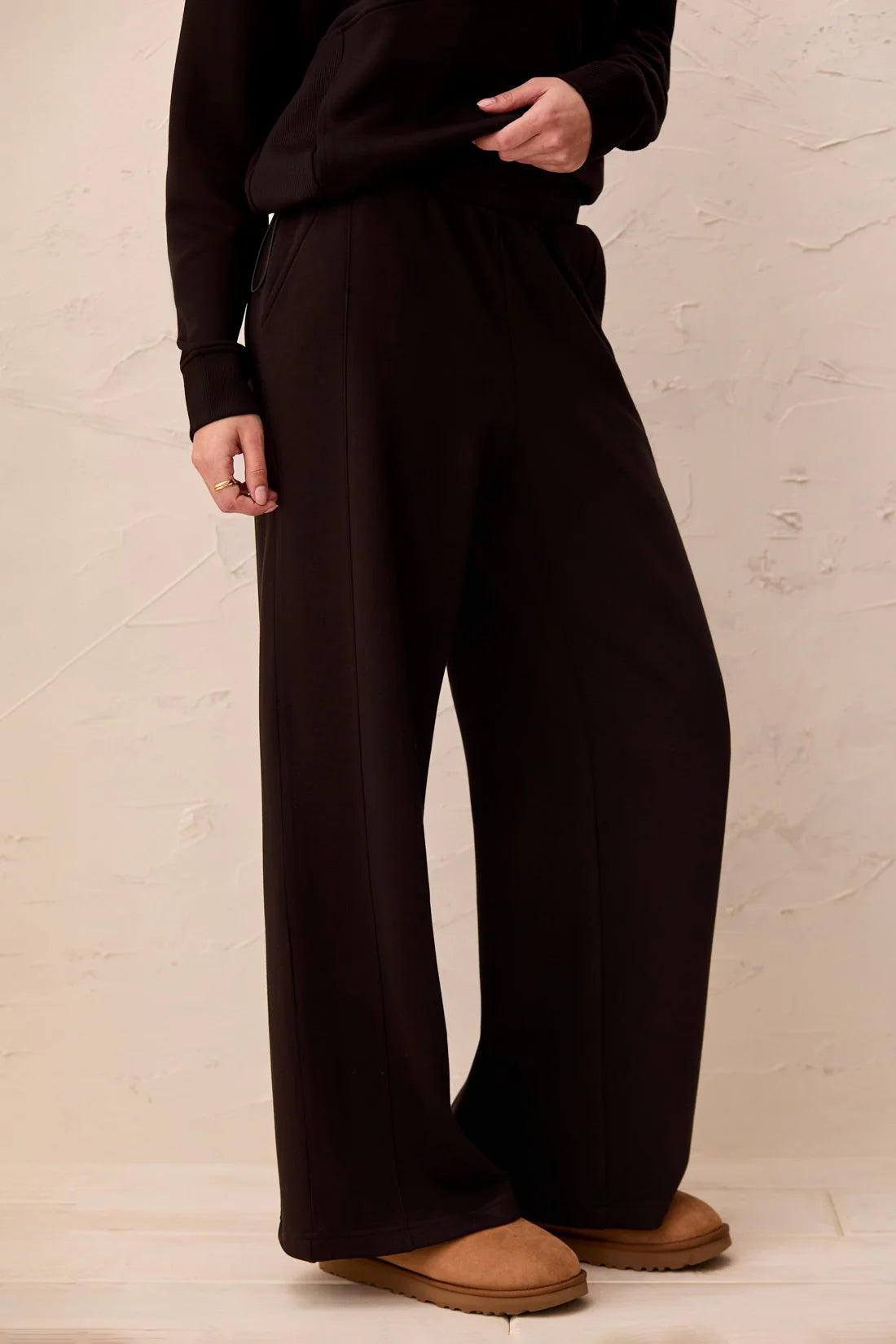 A person wearing dark brown wide-leg lounge pants with a matching dark top, standing against a textured beige wall.