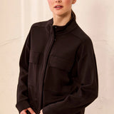 A woman wearing a dark brown zip-up jacket with a mock neck and flap chest pockets, paired with matching dark brown pants, set against a textured off-white wall.