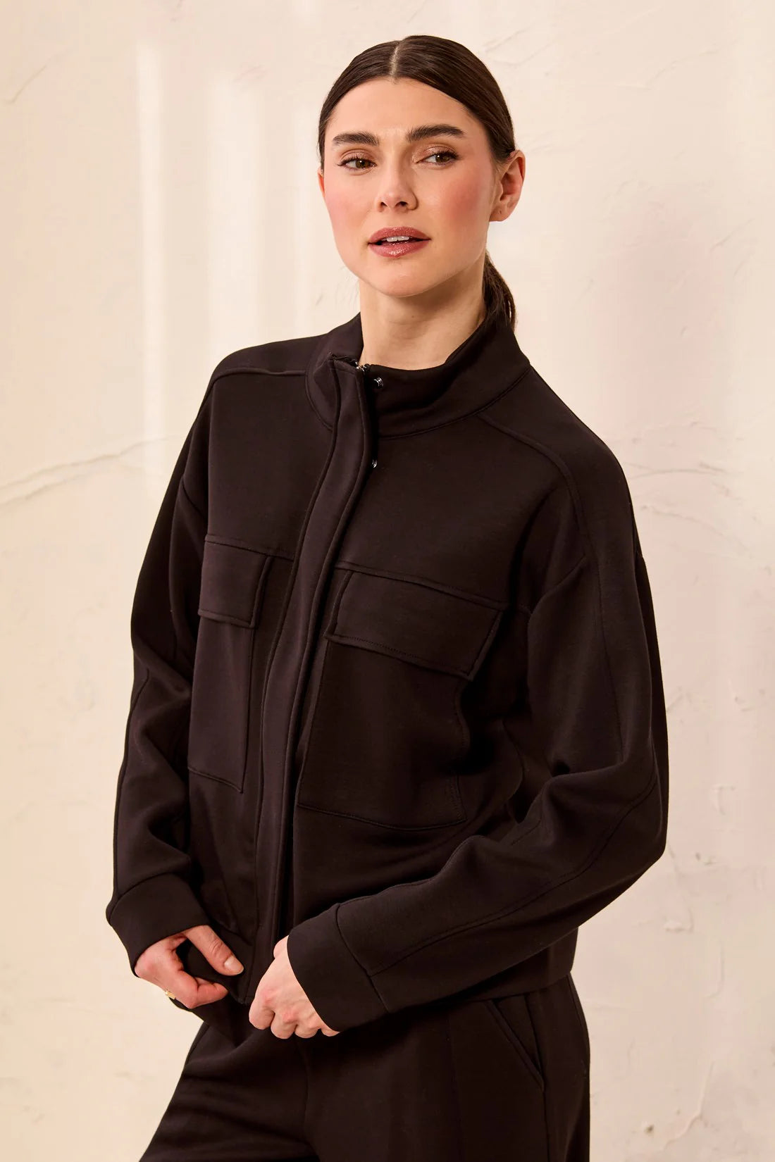 A woman wearing a dark brown zip-up jacket with a mock neck and flap chest pockets, paired with matching dark brown pants, set against a textured off-white wall.