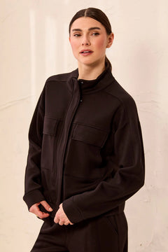 A woman wearing a dark brown zip-up jacket with a mock neck and flap chest pockets, paired with matching dark brown pants, set against a textured off-white wall.