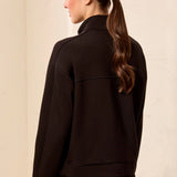 A woman shown from the back wearing a black cropped jacket with a mock neck and long sleeves, paired with black bottoms, against a light beige textured wall.